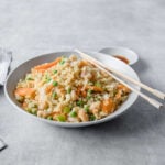 A shallow bowl filled with Shrimp Fried Cauliflower Rice, accented with carrots, peas, and green onions with chopsticks and side of extra coconut aminos.