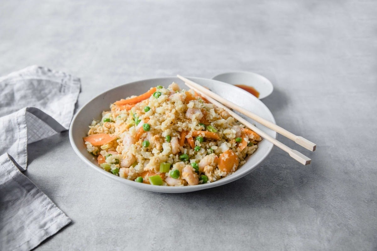 A shallow bowl filled with Shrimp Fried Cauliflower Rice, accented with carrots, peas, and green onions with chopsticks and side of extra coconut aminos.