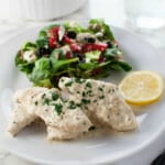 a white plate with creamy chicken, lemon slices and a mixed green salad.