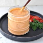 A jar of roasted red pepper aioli, served with a spoon. Roasted peppers and fresh parsley on the side as a garnish.