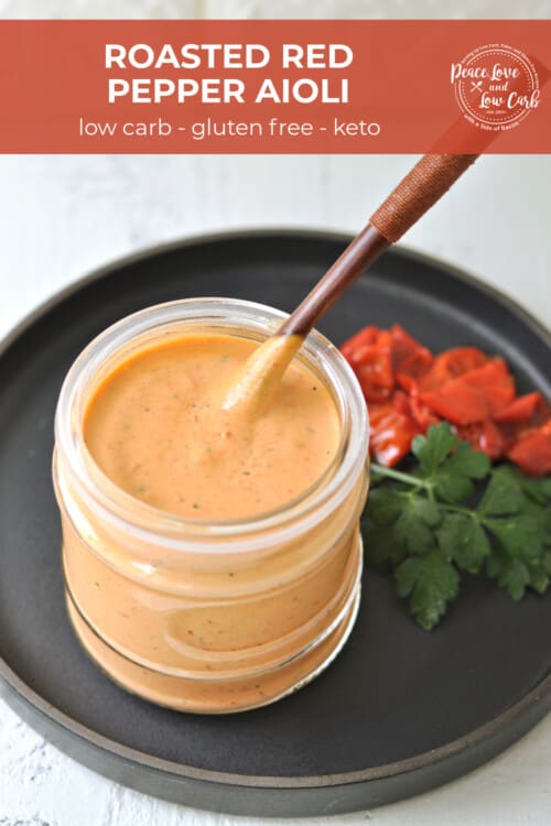 A jar of roasted red pepper aioli, served with a spoon. Roasted peppers and fresh parsley on the side as a garnish.