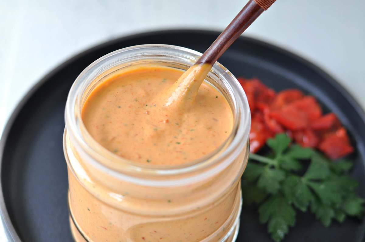 a jar of roasted red pepper aioli, served with a spoon. Roasted peppers and fresh parsley on the side as a garnish