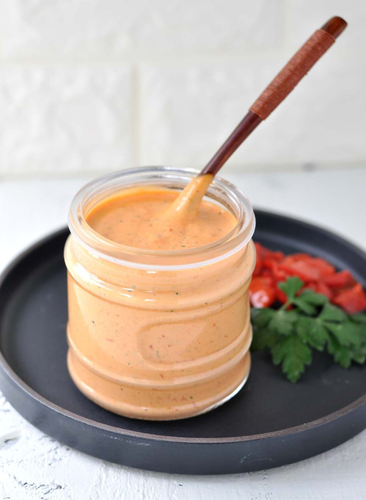 A jar of roasted red pepper aioli, served with a spoon. Roasted peppers and fresh parsley on the side as a garnish.