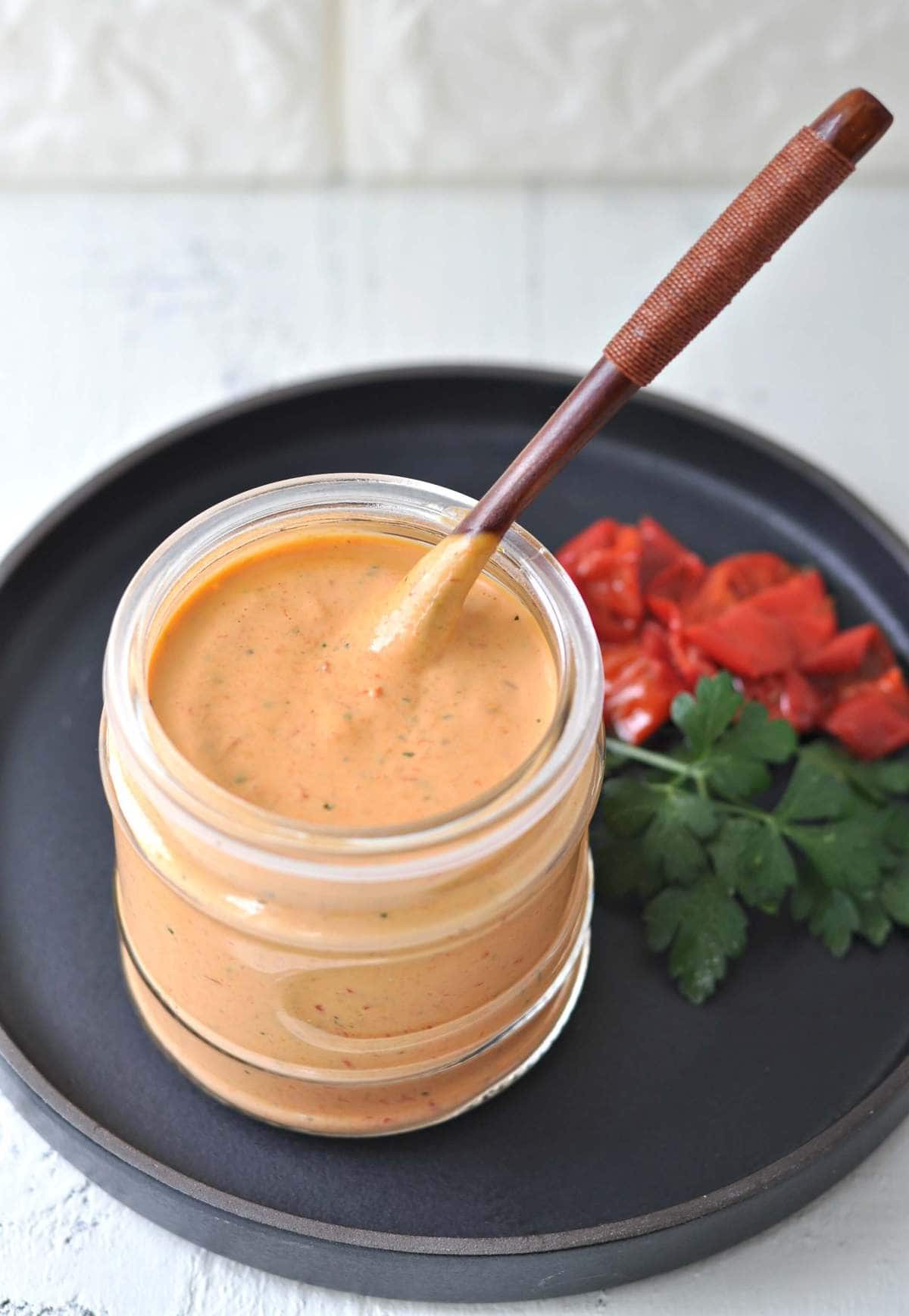 a jar of roasted red pepper aioli, served with a spoon. Roasted peppers and fresh parsley on the side as a garnish