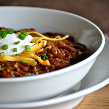 a closeup of a bowl of chili, topped with cheese, sour cream and green onions