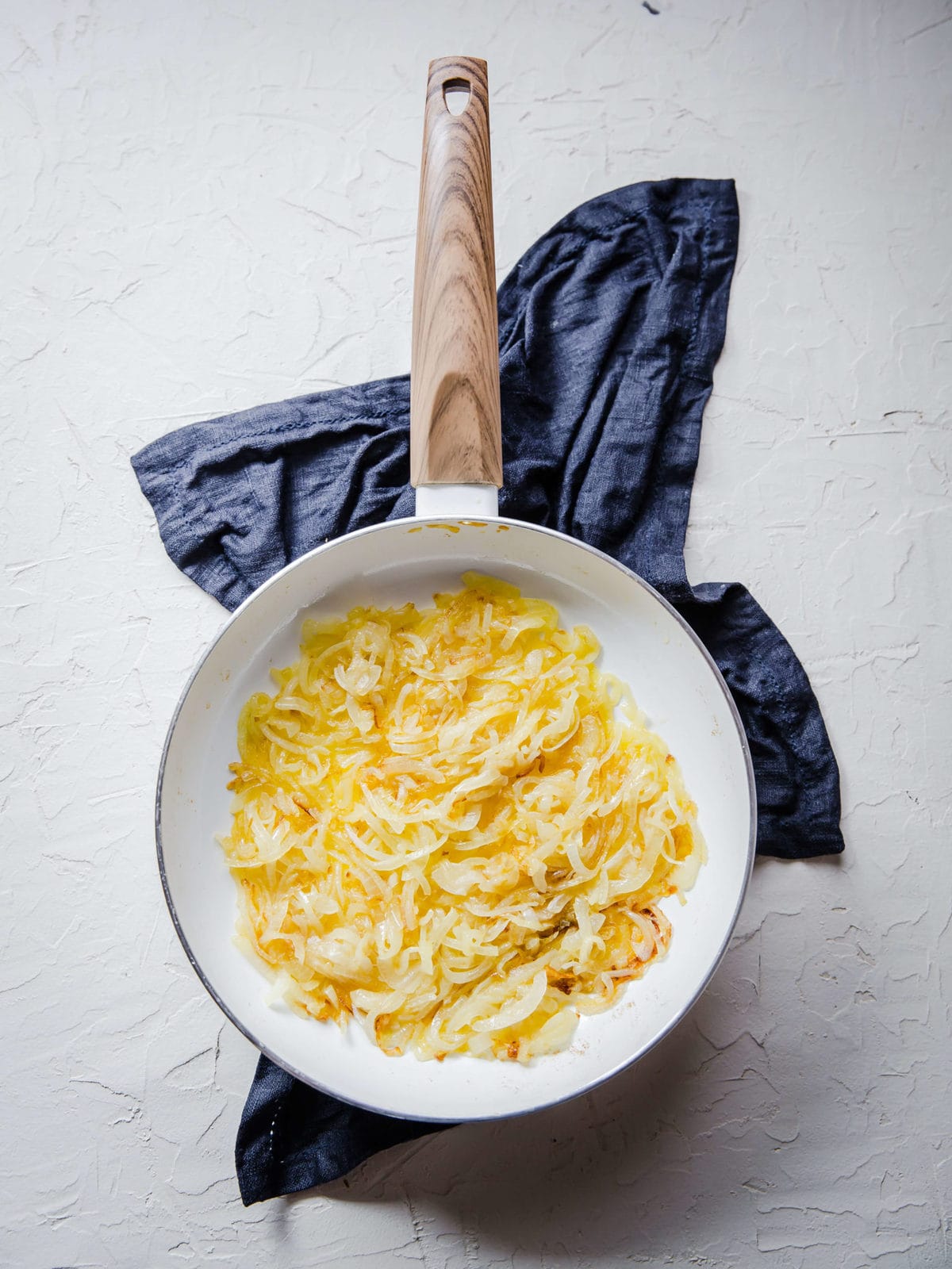 a white skillet full of onions sautéing in butter and sherry next to a blue hand towel.