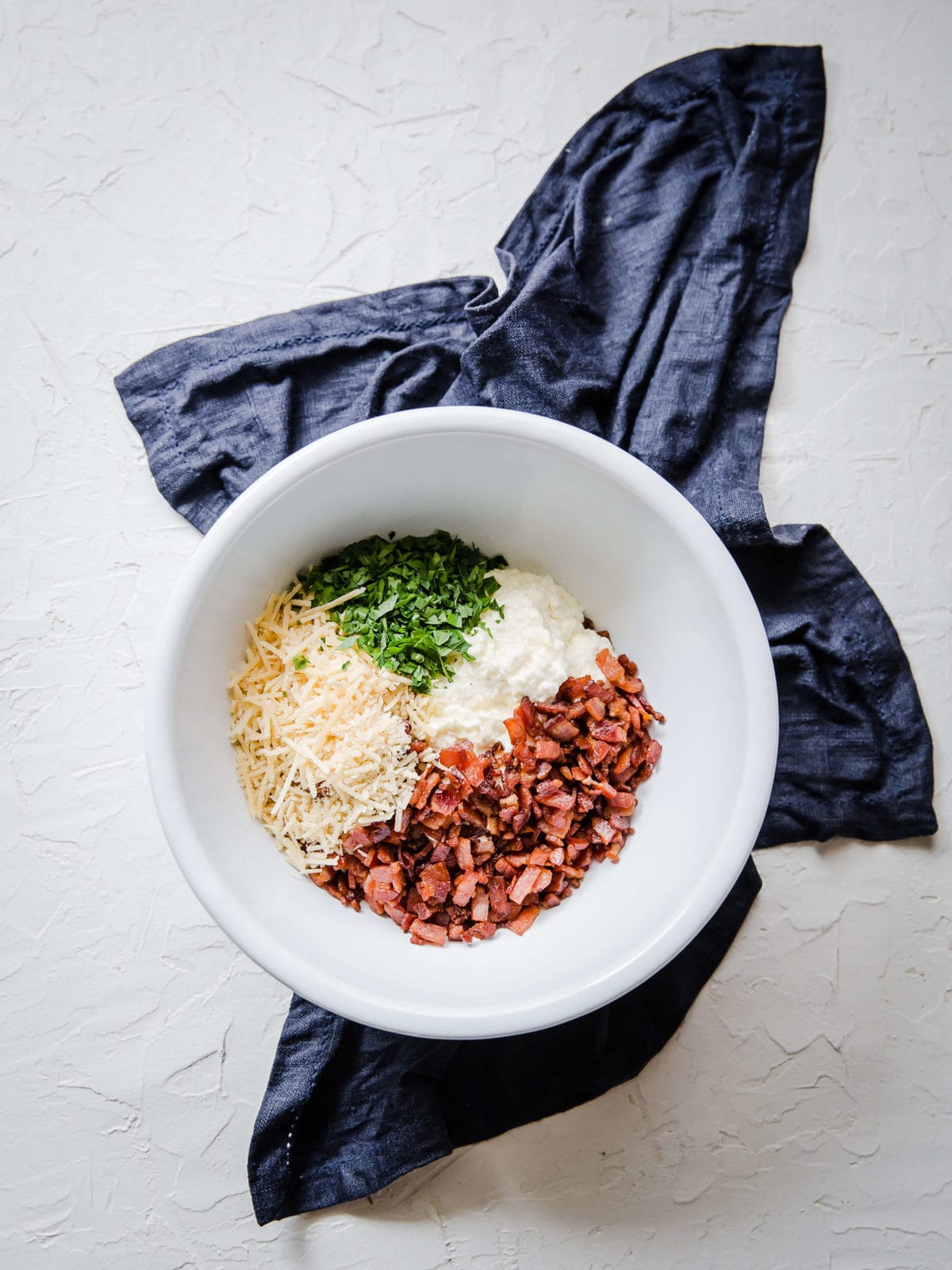 A white mixing bowl with cooked bacon, parmesan cheese, cream cheese, sour cream and fresh parsley.