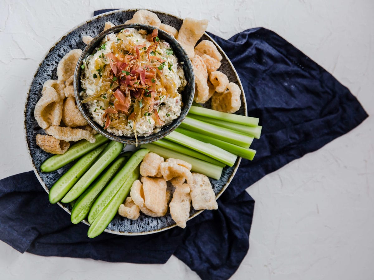 a gray bowl filled with caramelized onion dip and topped with bacon, served with pork rinds and fresh veggies.