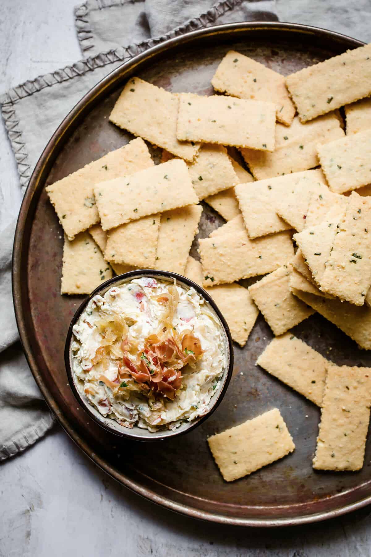 a gray bowl filled with caramelized onion dip and topped with bacon, served with garlic parmesan keto crackers.