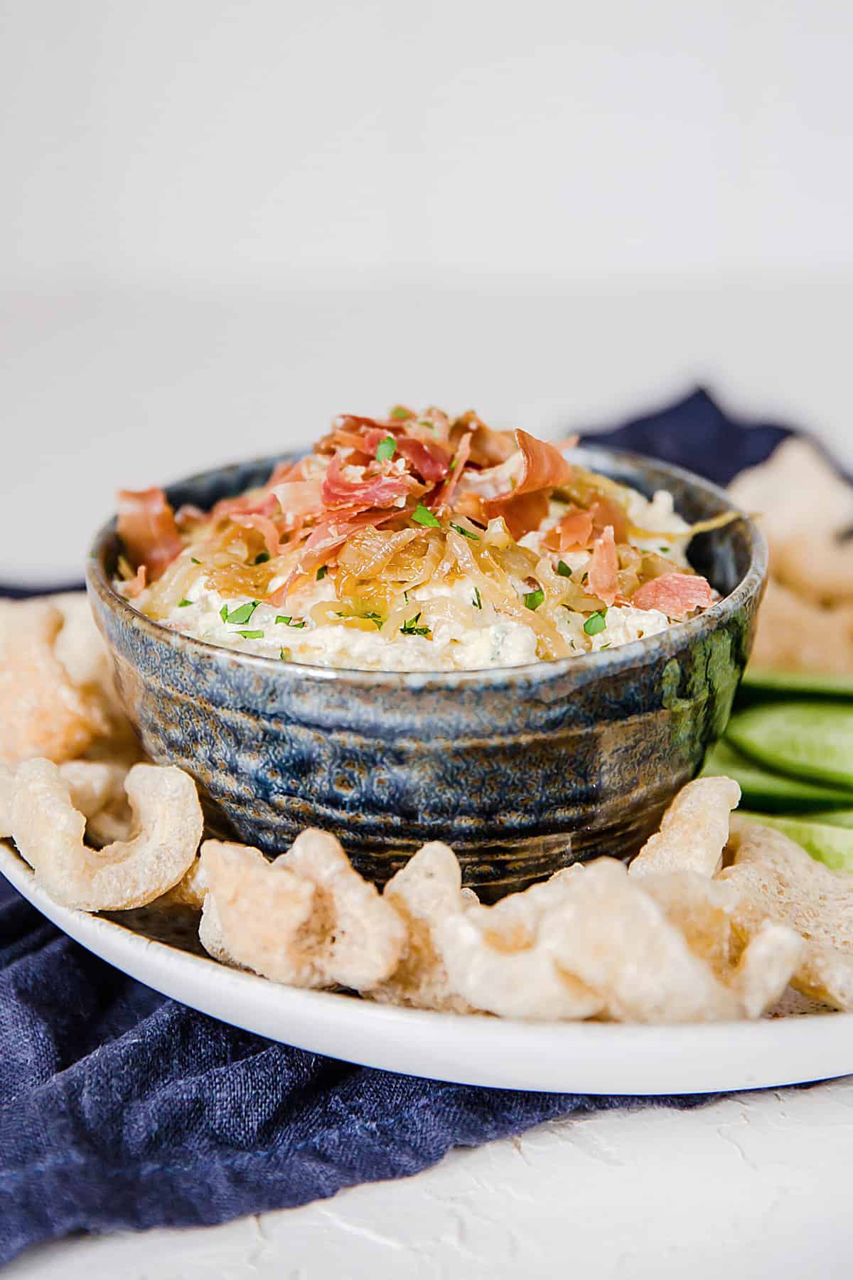 a gray bowl filled with caramelized onion dip and topped with bacon, served with pork rinds and fresh veggies.