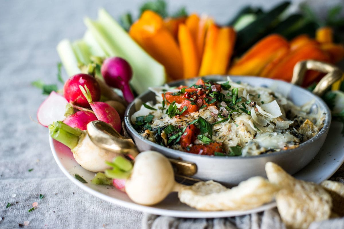 a metal bowl full of artichoke dip, topped with red peppers and parmesan cheese, surrounded by fresh vegetables and pork rinds