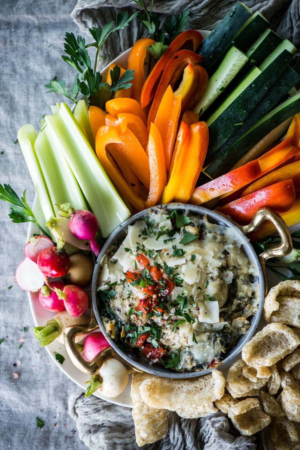 a metal bowl full of artichoke dip, topped with red peppers and parmesan cheese, surrounded by fresh vegetables and pork rinds