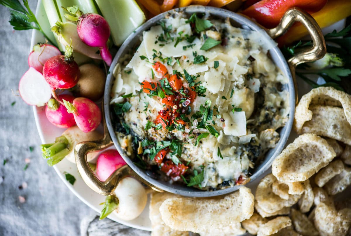 a metal bowl full of artichoke dip, topped with red peppers and parmesan cheese, surrounded by fresh vegetables and pork rinds