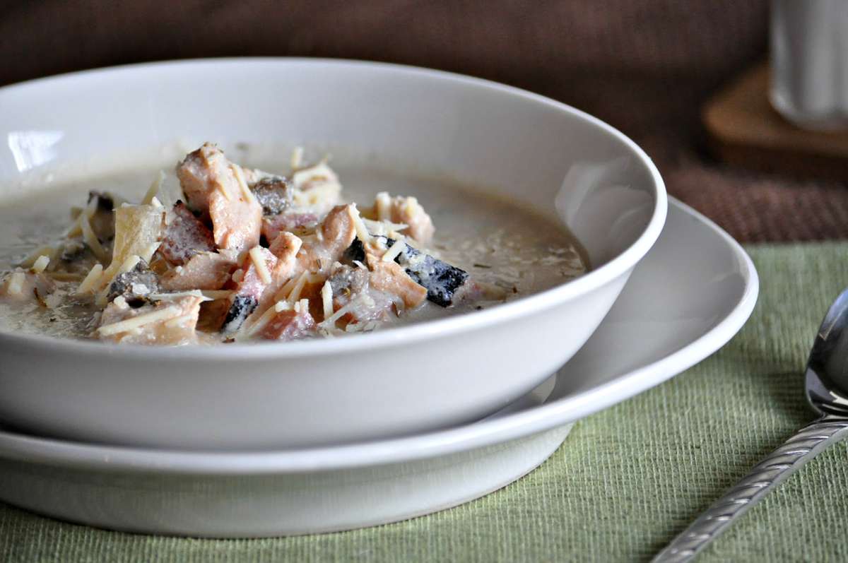 a white bowl, full of soup with chicken, ham, mushrooms, and cheese.