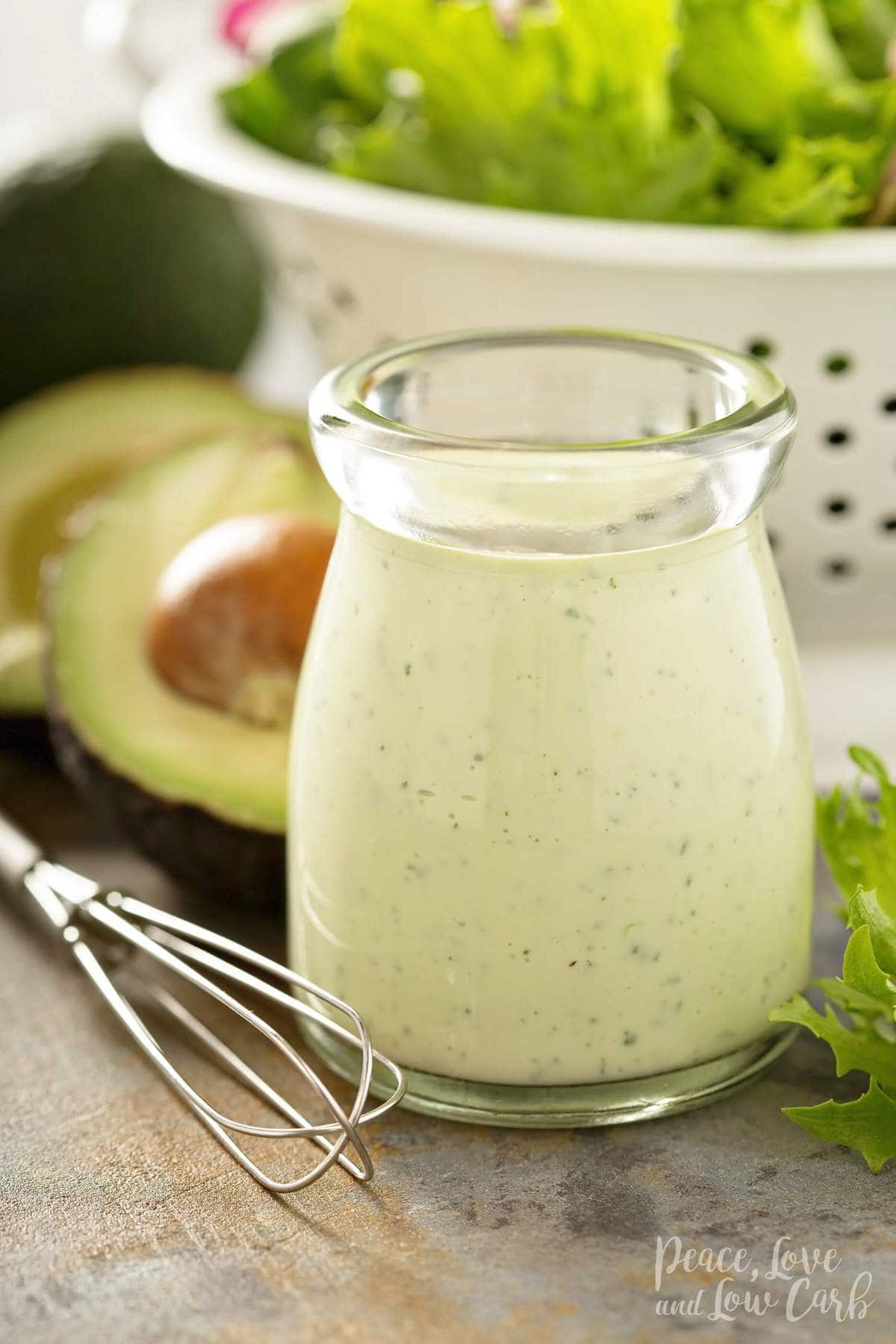 a jar of creamy salad dressing with lettuce and avocado behind it. 