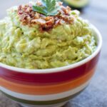 A red, orange, and white striped bowl filled to the brim with fresh, creamy guacamole, topped with crispy bacon and a cilantro leaf.