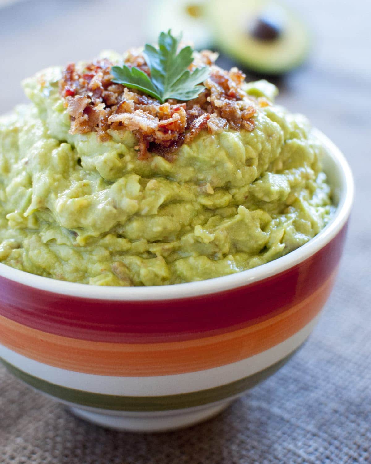 A red, orange, and white striped bowl filled to the brim with fresh, creamy guacamole, topped with crispy bacon and a cilantro leaf.