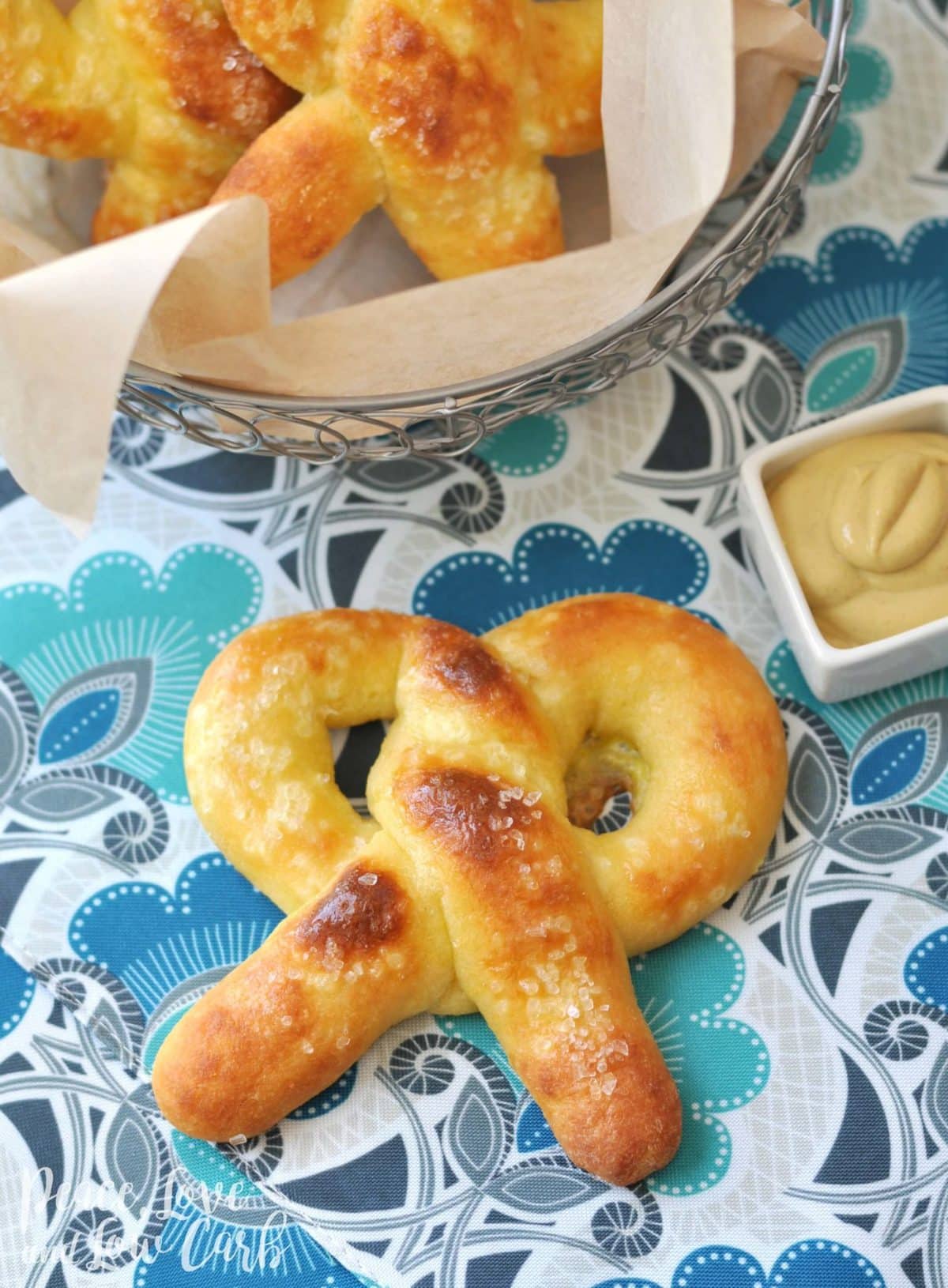 On a gray countertop with a white patterned backsplash, a metal basked with parchment paper loaded with keto soft pretzels sits to the left side of the frame on top of an aqua, gray, black, and blue tablecloth. At the center of the photo sits a golden brown keto soft pretzel with a small white square sauce dish full of mustard.