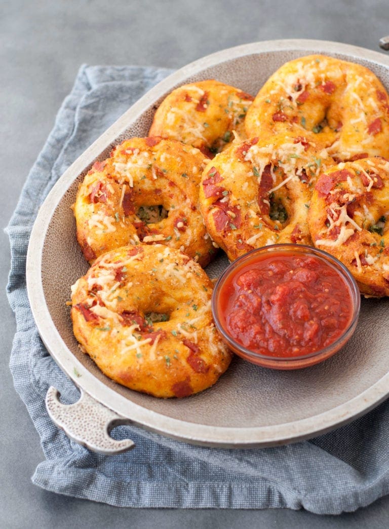 Overhead shot of 6 keto pizza bagels sitting on a light gray metal plate with a glass sauce bowl full of marinara. The plate sits on light gray fabric napkins, on a dark gray countertop.
