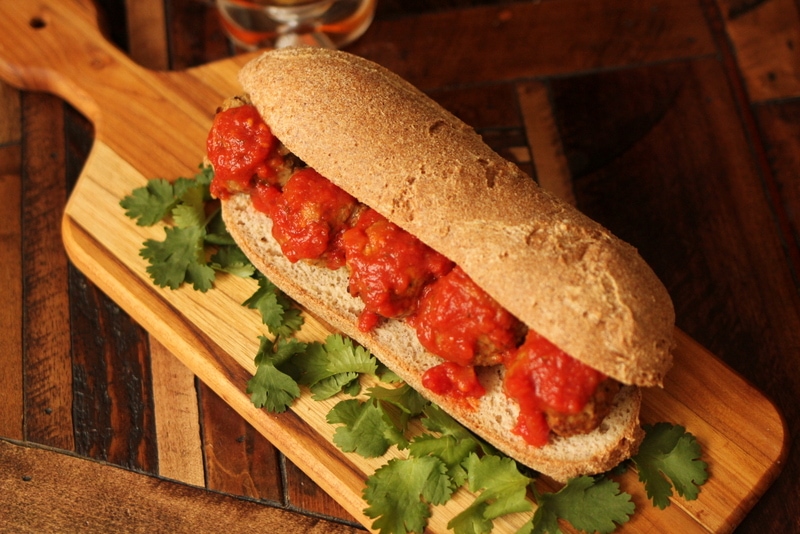 A healthy sub roll is sliced down the middle and loaded with meatballs and marinara. The sub sits on a bed of fresh herbs on a wooden cutting board, resting on a wooden countertop.
