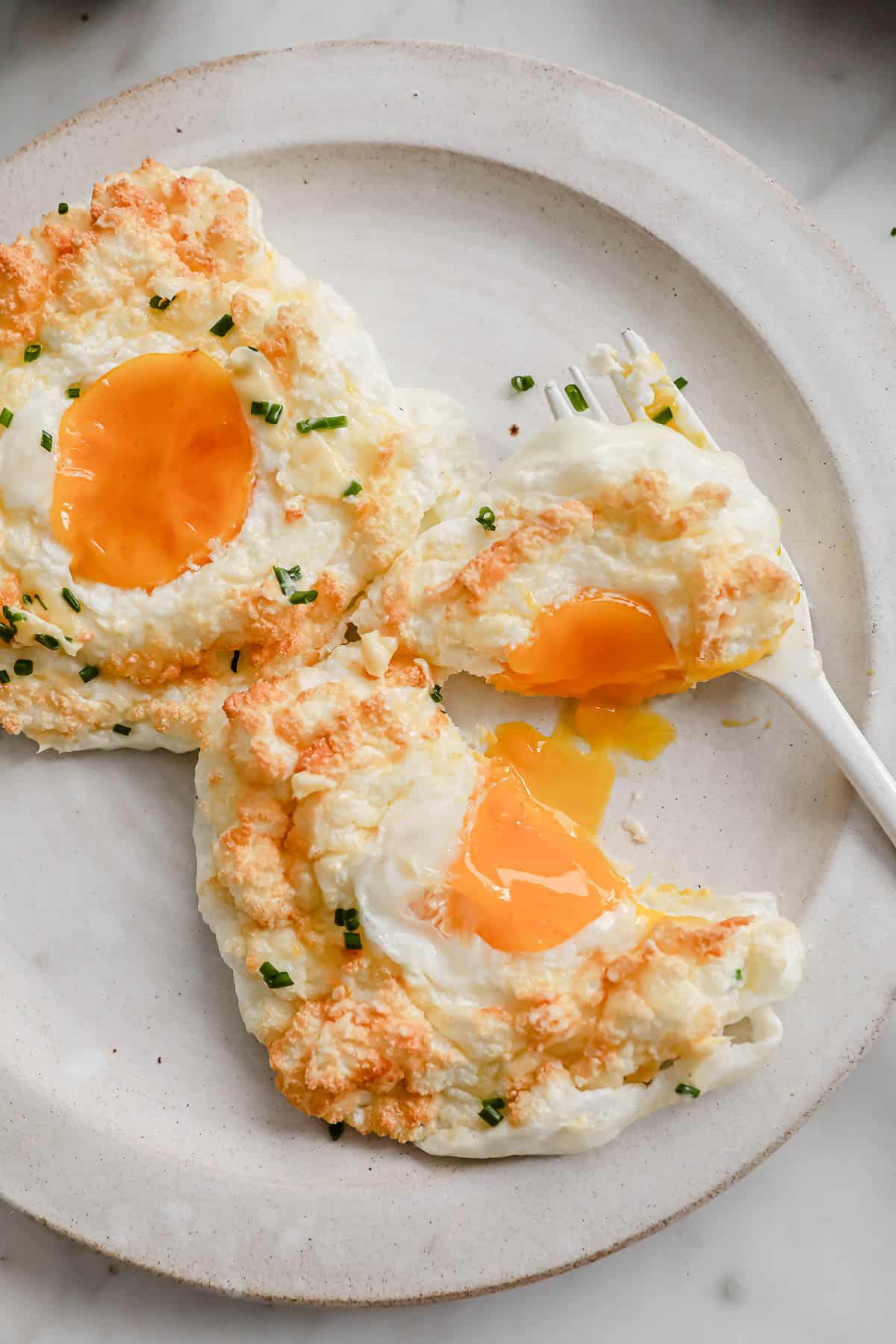 2 baked egg clouds, topped with chives, on a white ceramic plate with a fork