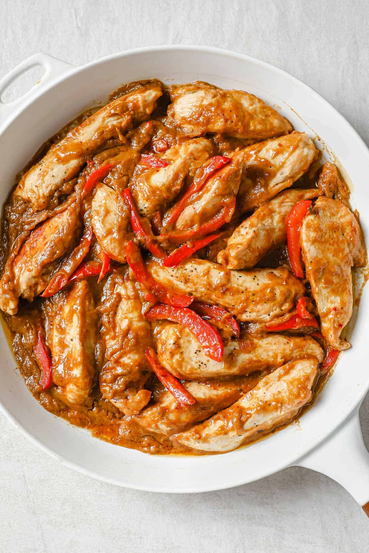 a white skillet with pan-seared chicken, red peppers, and a peanut butter sauce