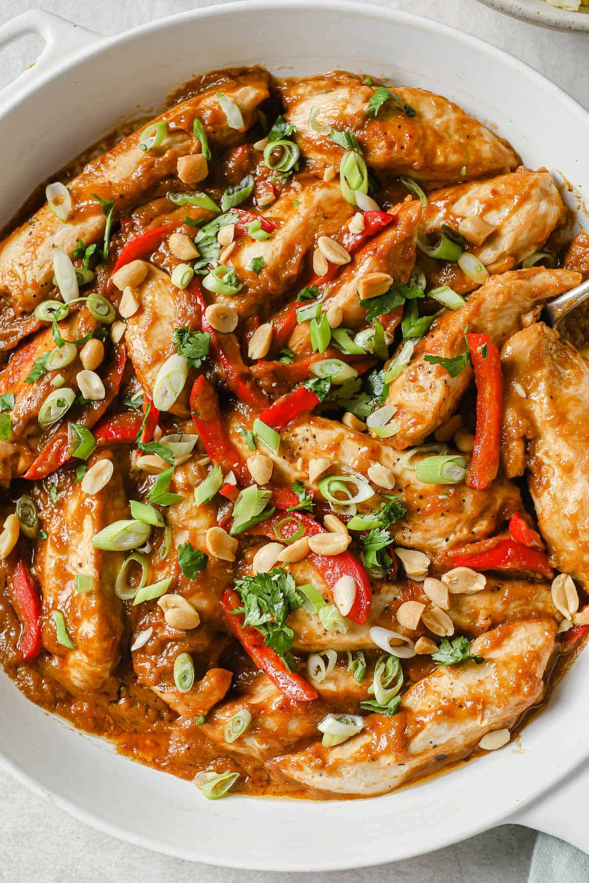 white skillet full of chicken, with a peanut butter sauce, red bell peppers, green onions and cilantro