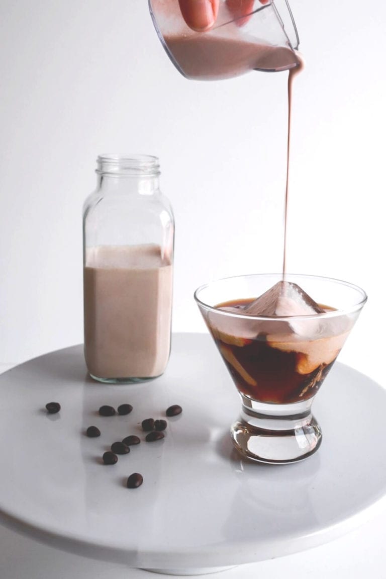 A mini martini glass with a large block of ice, partially filled with coffee is drizzled with Irish Cream mix. The martini glass sits on a marble cake stand, sprinkled with coffee beans, and a large glass bottle of Irish Cream sits towards the back of the photo, all framed by a white backdrop.