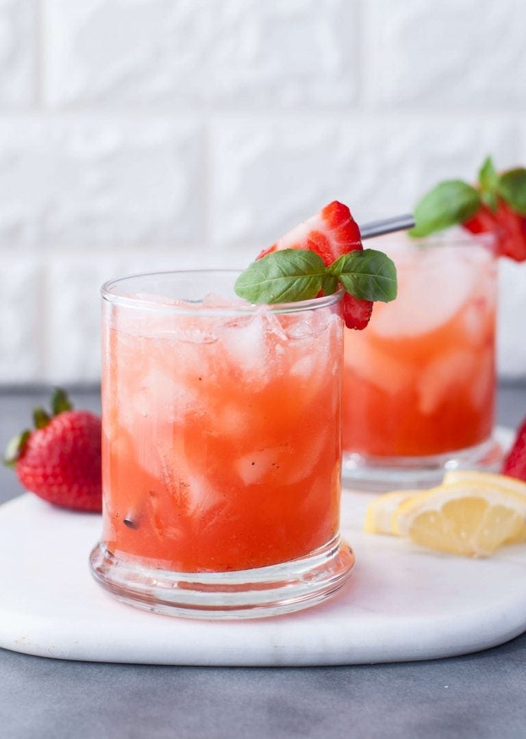 A lowball glass filled with ice cubes, a metal straw, and Strawberry Basil Bourbon Smash, garnished with fresh strawberry and mint, sits on a marble slab on a gray countertop with a white brick background. On the marble slab sits another whole fresh strawberry and lemon wedges, with a second drink in the background.