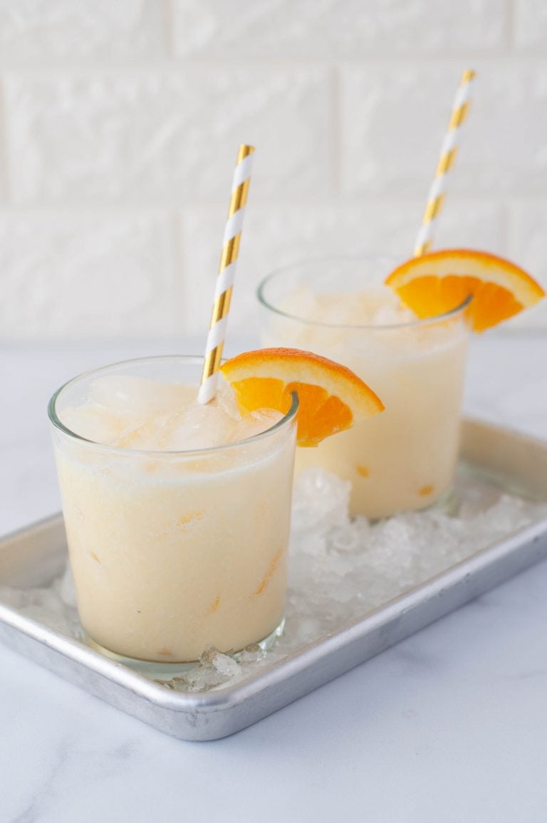 A small rectangular aluminum baking sheet sits on a white marble countertop. The baking sheet is filled with ice and two Orange Creamsicle Cocktails, each in a lowball glass with ice, an orange wedge garnish, and a white and gold striped paper straw.
