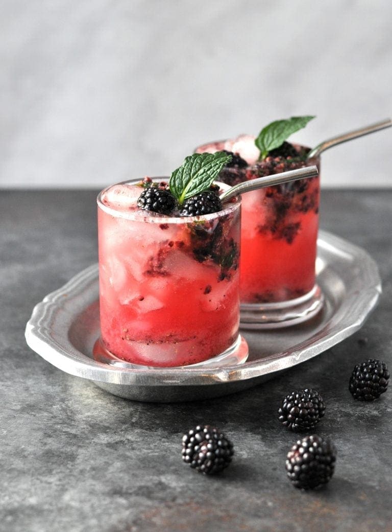 A silver serving tray sits on a dark gray countertop with a light gray background. On the serving tray, there are two lowball glasses filled with ice, fresh muddled blackberries, and Black Beauty cocktail, garnished with fresh mint and a metal straw. A few fresh blackberries are littered on the countertop.