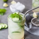 A mason jar filled with low carb mojito, ice, a metal straw, fresh mint, and lime slices sits on top of a wooden coaster on a gray countertop with a white brick background. In the background, extra garnish is scattered on the countertop along with a bowl of ice, a gray cloth napkin with a martini shaker lid, and an additional drink.