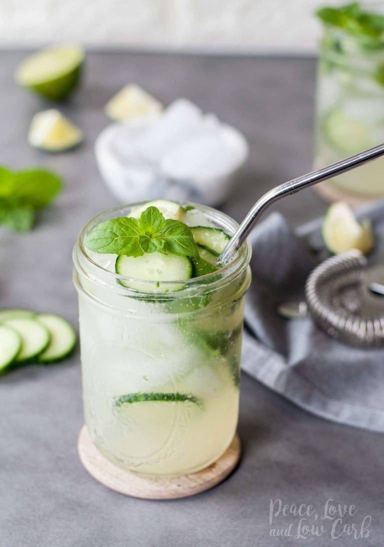 A mason jar filled with low carb mojito, ice, a metal straw, fresh mint, and lime slices sits on top of a wooden coaster on a gray countertop with a white brick background. In the background, extra garnish is scattered on the countertop along with a bowl of ice, a gray cloth napkin with a martini shaker lid, and an additional drink.