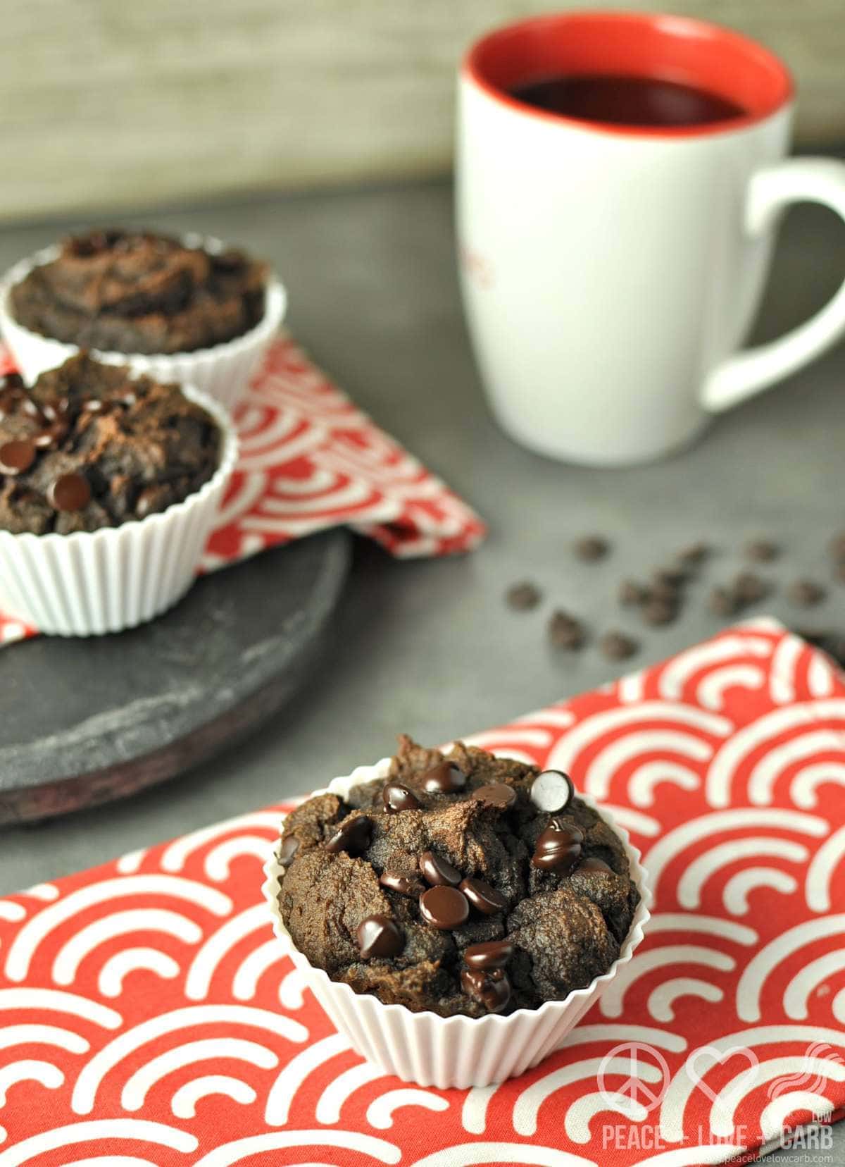 a chocolate muffin on a red and white striped towel, with more muffins and a mug of coffee in the background. 