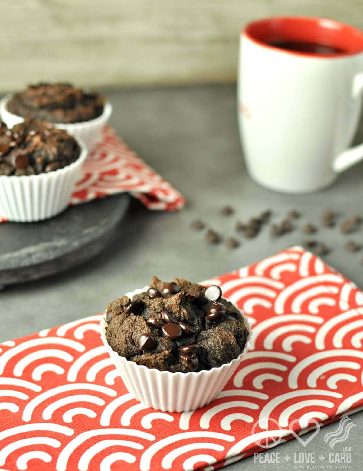 a chocolate muffin on a red and white striped towel, with more muffins and a mug of coffee in the background. 
