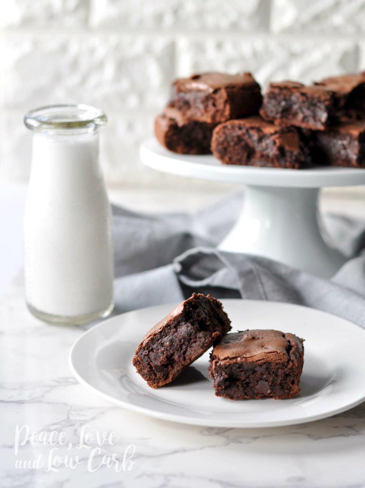 A plate with 2 brownies, next to a glass of milk, with a cake platter of brownies behind it. 