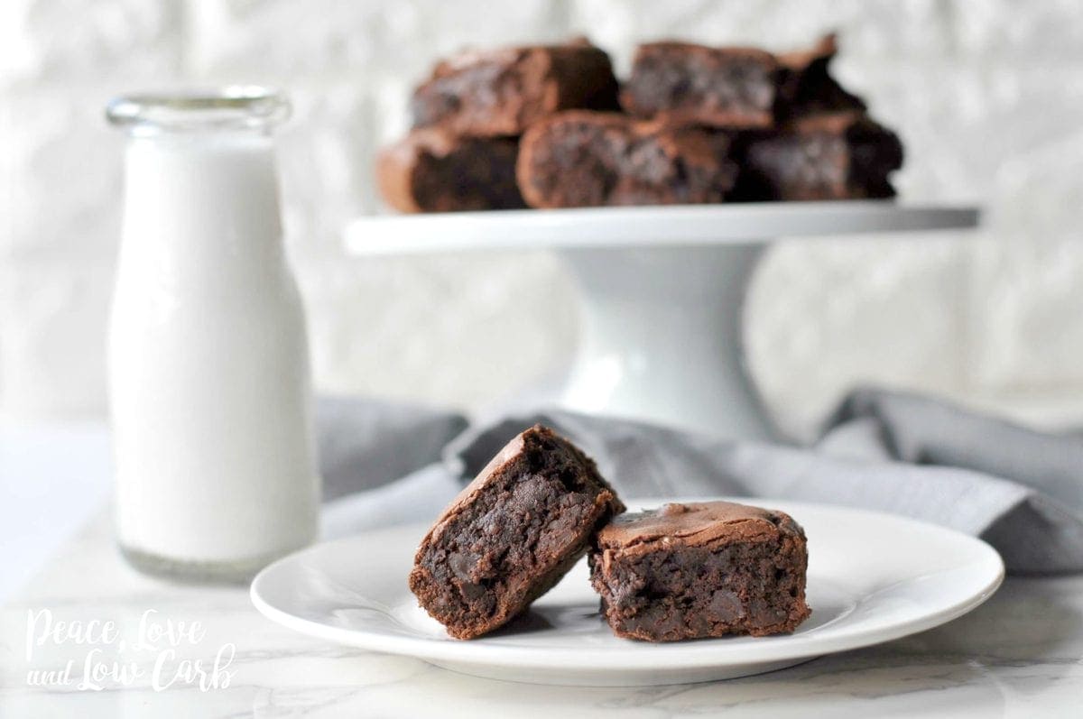 A plate with 2 brownies, next to a glass of milk, with a cake platter of brownies behind it. 