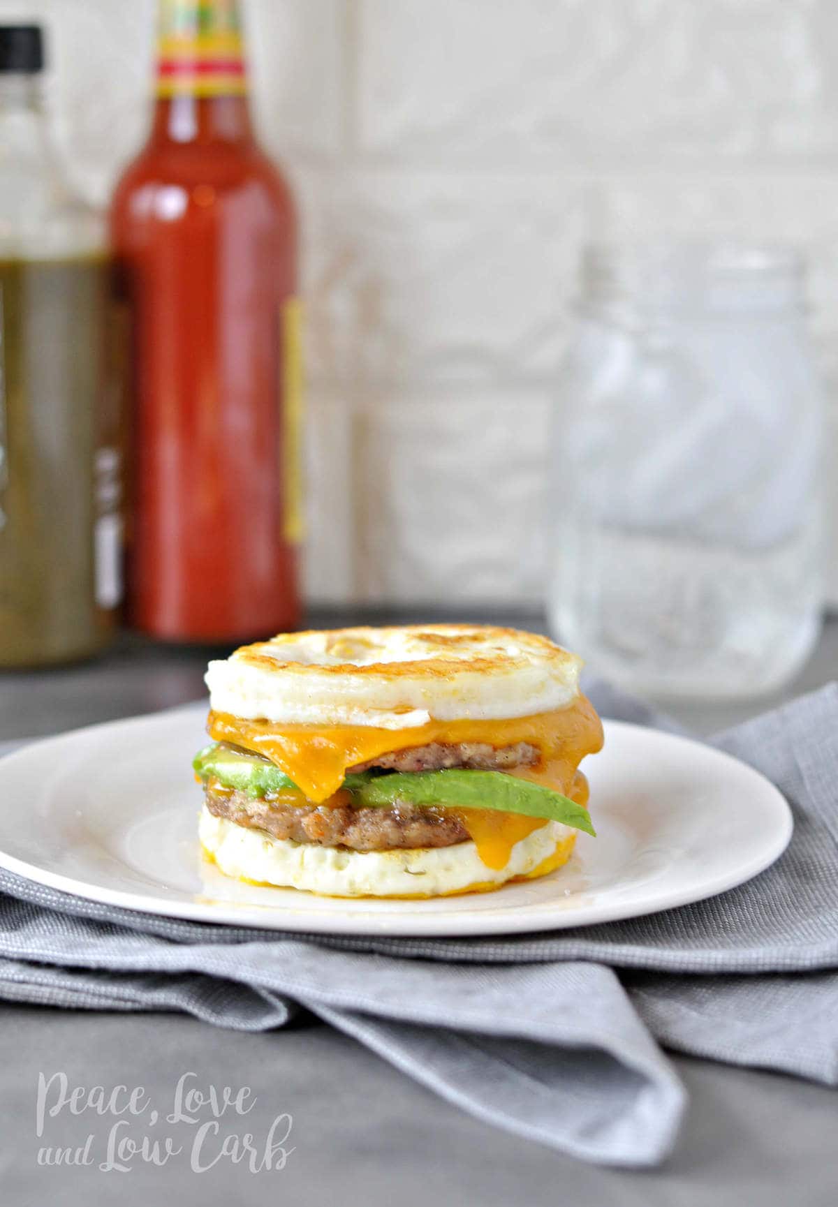 

an egg, sausage, cheese and avocado breakfast sandwich on a white plate, with a gray towel. 
