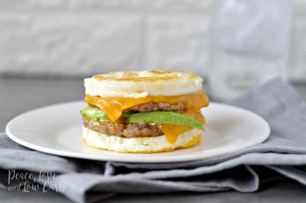 an egg, sausage, cheese and avocado breakfast sandwich on a white plate, with a gray towel. 