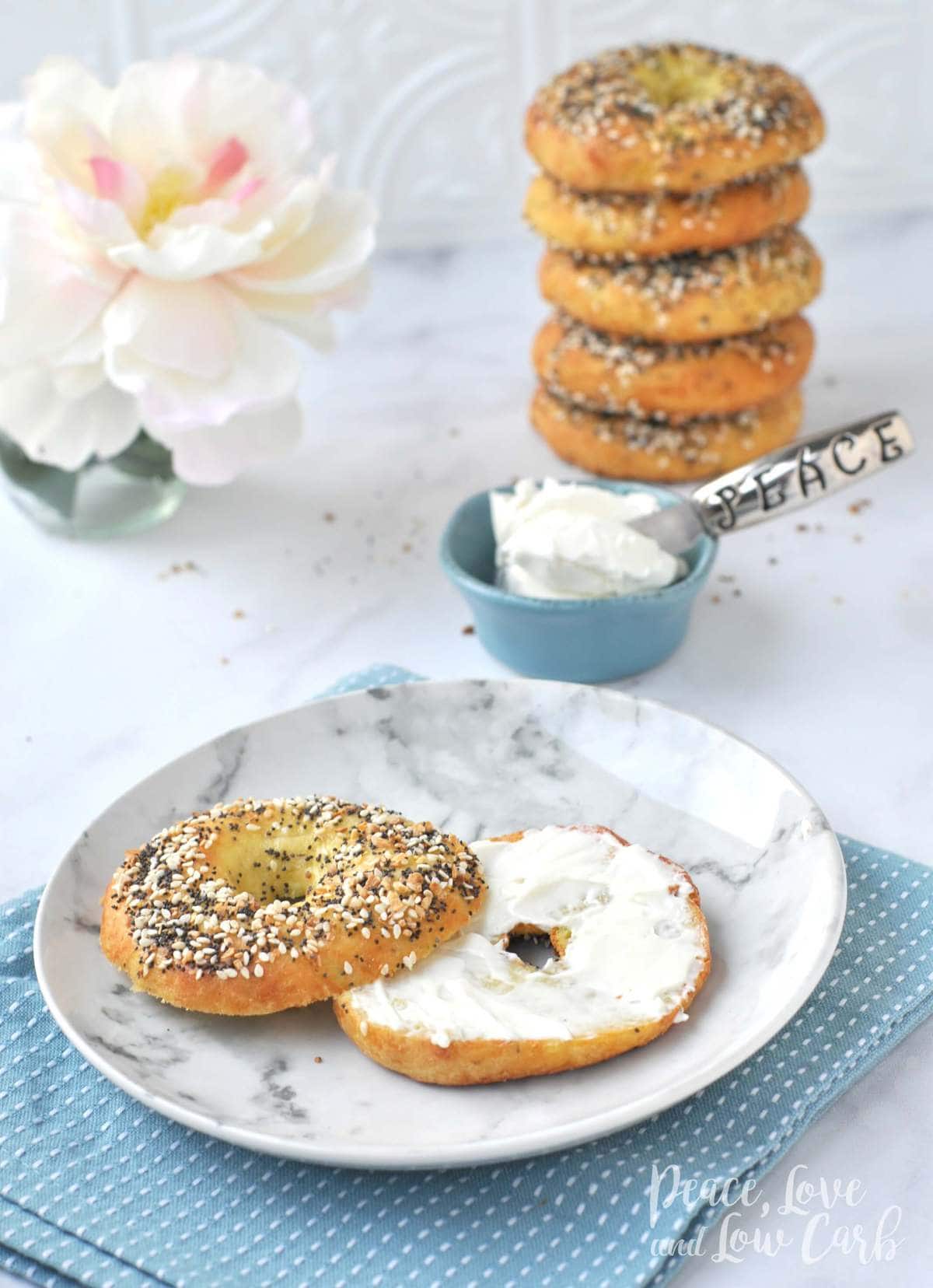 A low carb everything bagel with cream cheese on a marble plate. Behind it is a dish of cream cheese and a stack of bagels