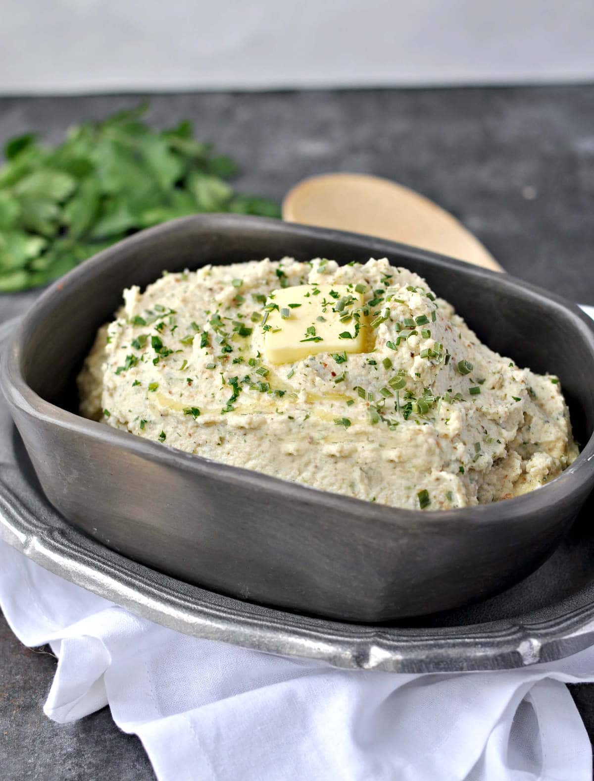 a dish of mashed cauliflower, garnished with butter and parsley, served with a wood spoon