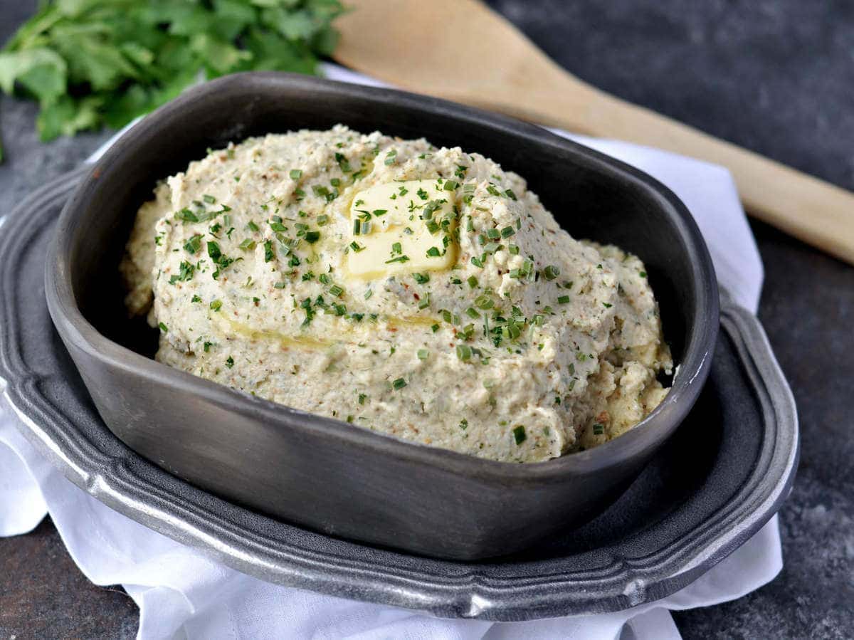 a dish of mashed cauliflower, garnished with butter and parsley, served with a wood spoon