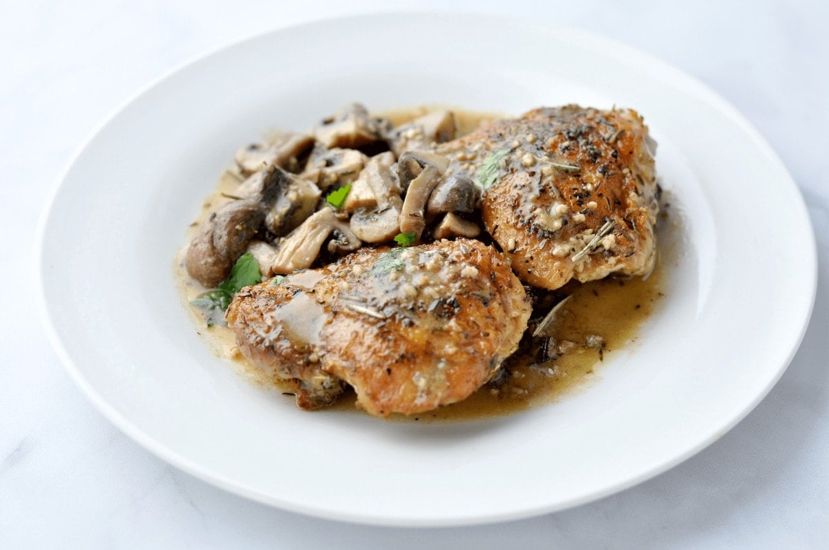 two crispy chicken thighs on a white plate, topped with a mushroom herb sauce