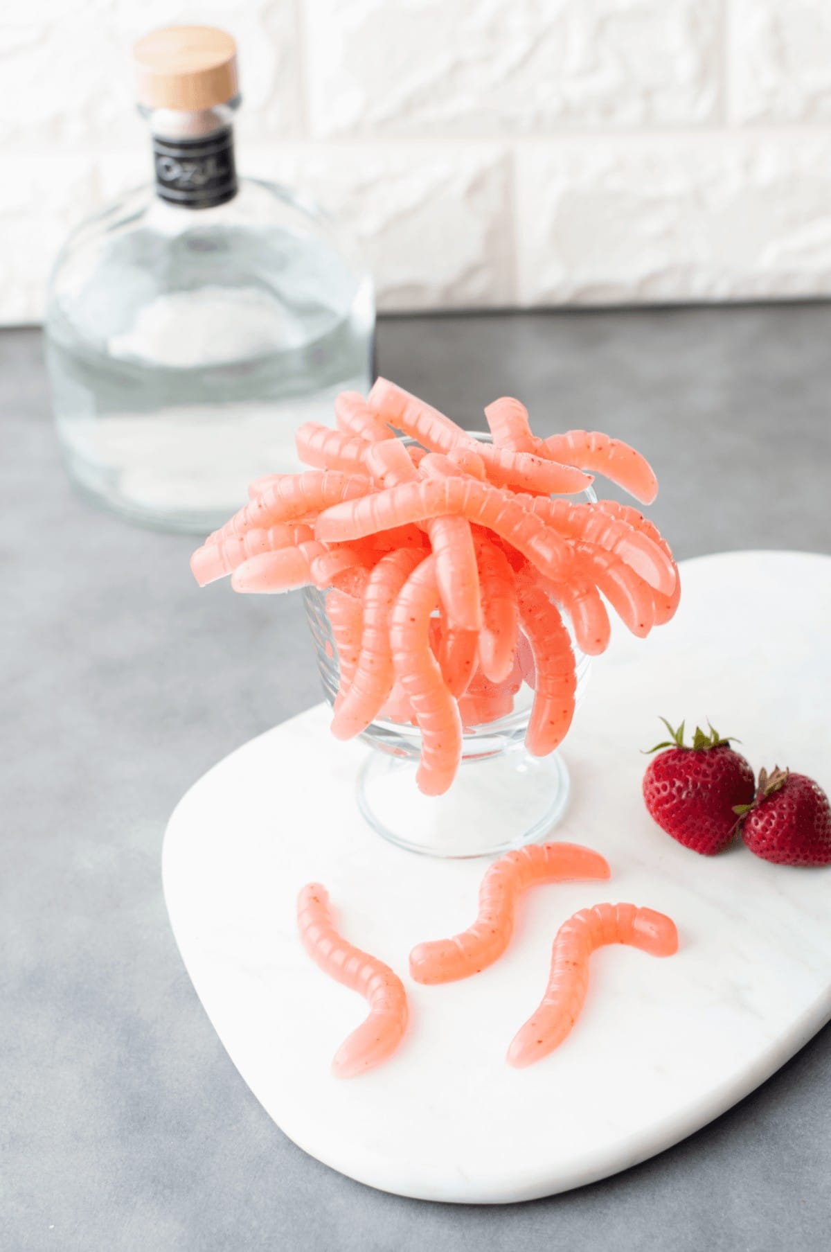 A clear glass full of strawberry daiquiri flavored gummy worms, with a bottle of tequila in the background. 