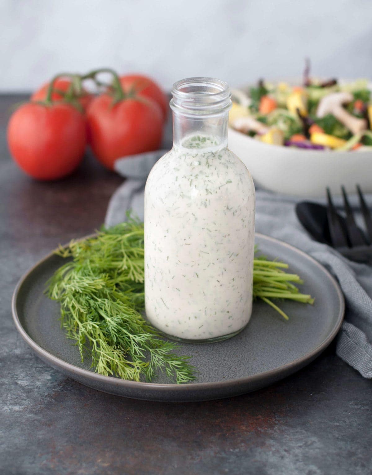 a jar of homemade ranch dressing with fresh dill next to it and a salad and tomatoes in the background