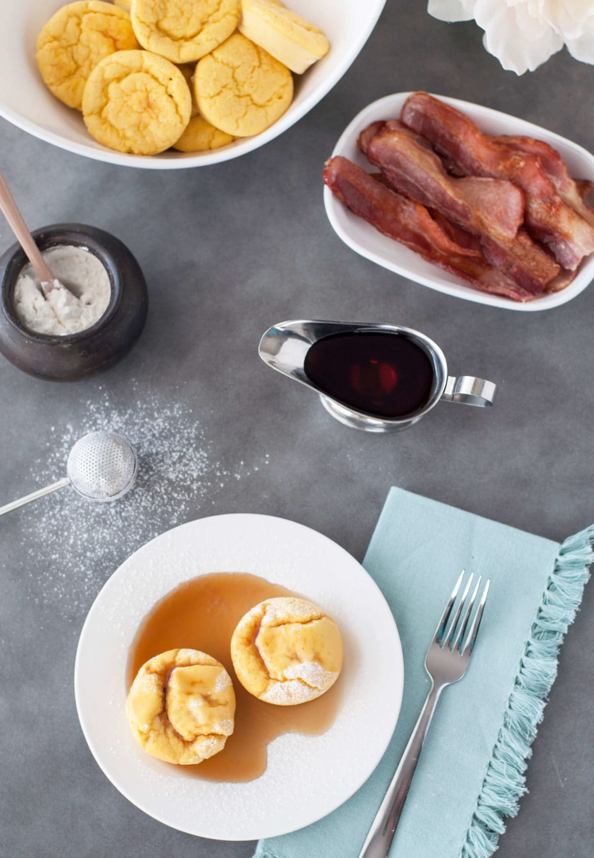 2 French toast egg puffs on a white plate, with syrup and bacon in the background