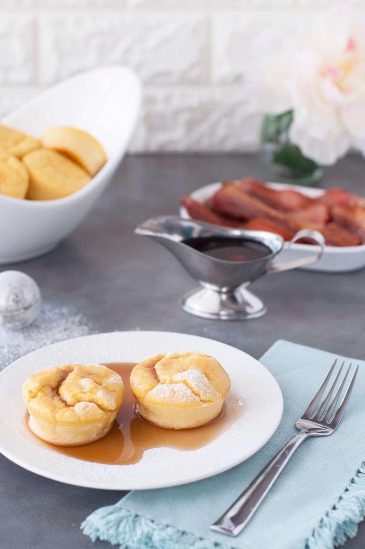 2 French toast egg puffs on a white plate, with syrup and bacon in the background