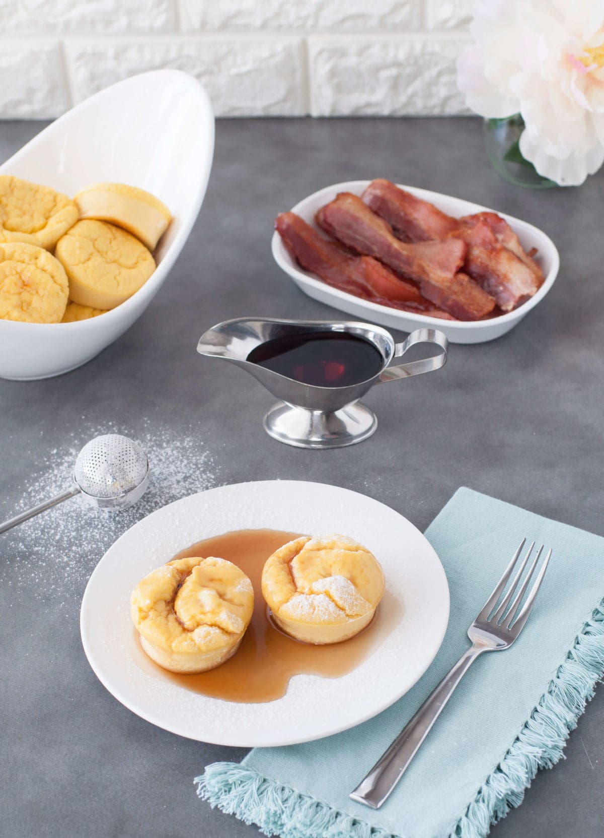 2 French toast egg puffs on a white plate, with syrup and bacon in the background