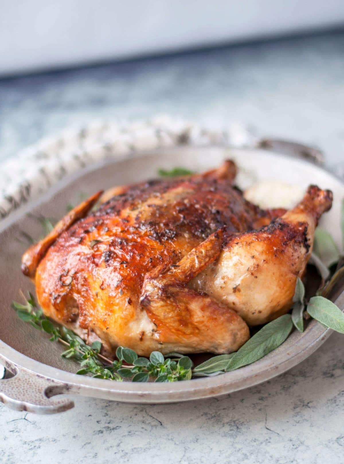 a whole roasted chicken with crispy skin on a metal platter, garnished with garlic and fresh herbs