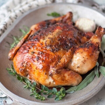 a whole roasted chicken with crispy skin on a metal platter, garnished with garlic and fresh herbs